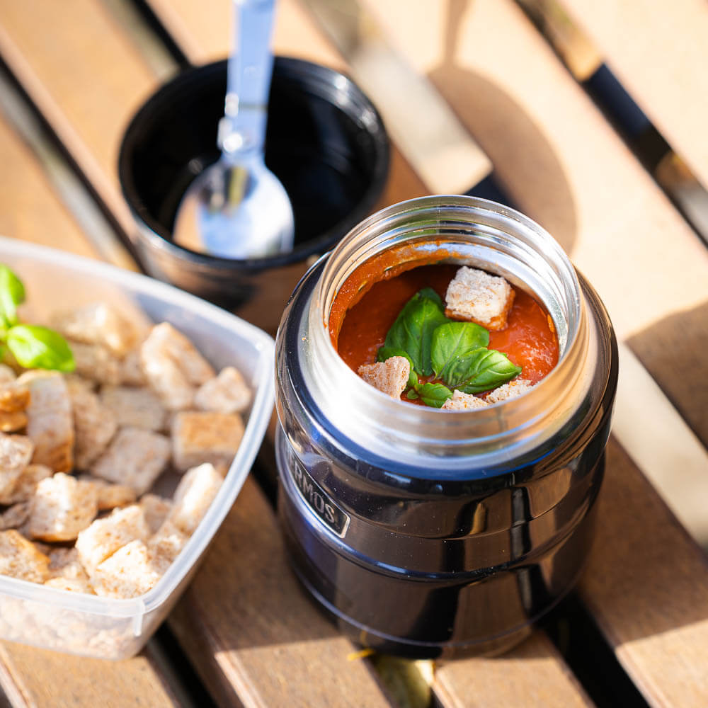 Eine geöffnete Thermoskanne zeigt eine frische Tomatensuppe mit Basilikum und Croutons. Neben der Thermoskanne steht ein Behälter mit zusätzlichen Croutons und ein Löffel.