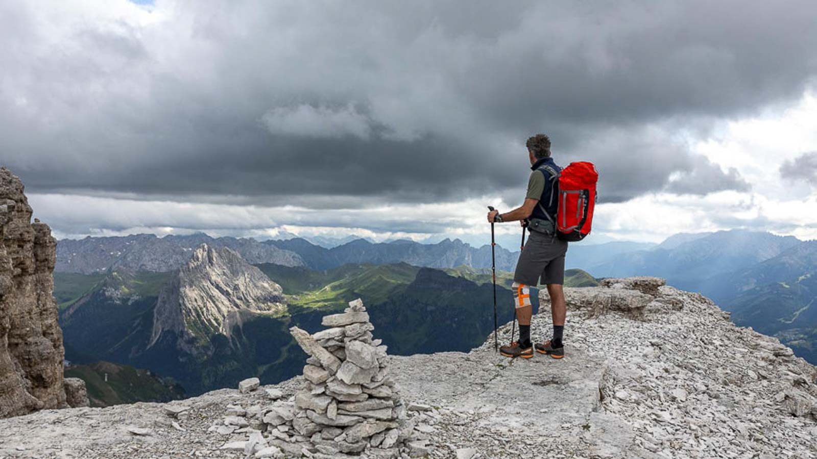 Eine Person mit einem roten Rucksack steht auf einem Felsvorsprung in den Dolomiten und blickt in die Ferne. Der Himmel ist bewölkt und es gibt eine Steinmännchen-Konstruktion im Vordergrund.