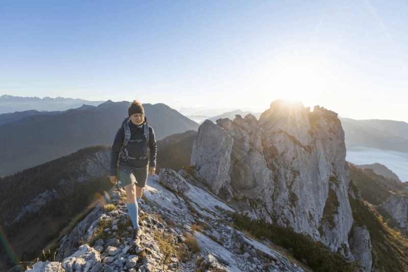 Eine Wanderin mit Outdoor Merino Compression Socks geht bei Sonnenaufgang auf einem schmalen Gebirgspfad. Diese Kompressionssocken bieten optimale Unterstützung und Komfort für anspruchsvolle Wanderungen.