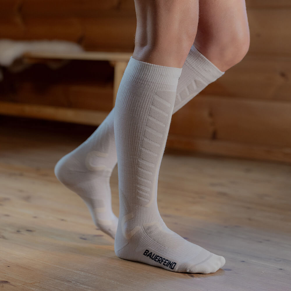 Nahaufnahme von weißen Kompressions-Skisocken, die bis unter die Knie reichen. Die Person steht auf einem Holzboden in einer Hütte. Die Socken bieten durch ihre gepolsterten Zonen zusätzlichen Komfort und Halt beim Skifahren.