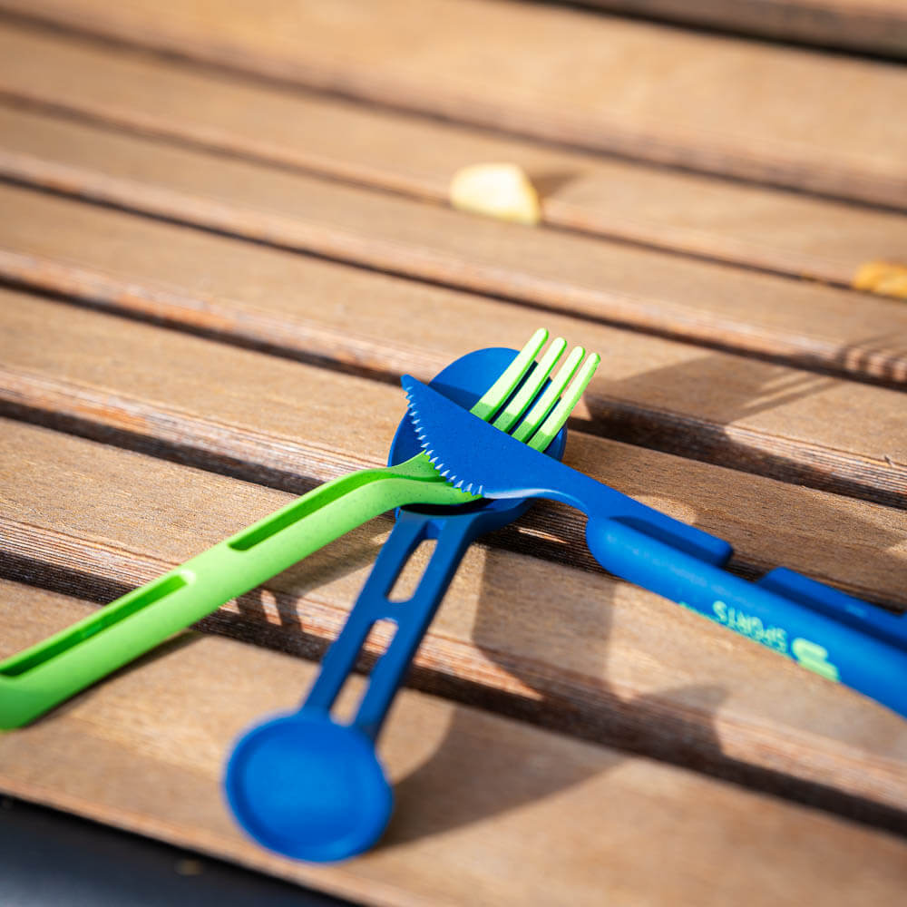 Ein grünes und ein blaues Besteck liegen kreuzweise auf einer Holzbank. Das Set ist ideal für Outdoor-Essen und platzsparendes Packen.