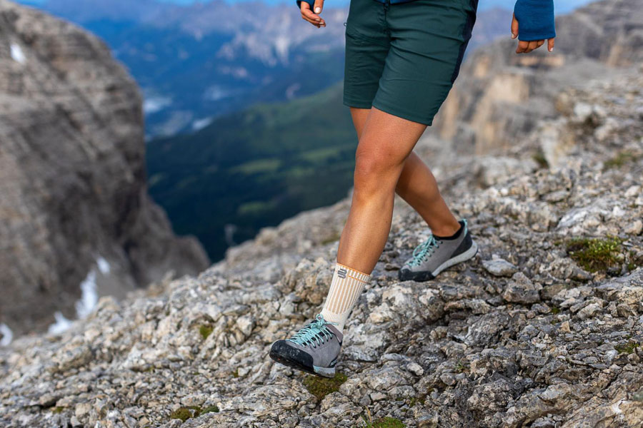 Ein Wanderer bewegt sich über felsiges Gelände mit Outdoor-Schuhen und einer orangefarbenen Sprunggelenksbandage. Im Hintergrund erstreckt sich eine weite Berglandschaft.