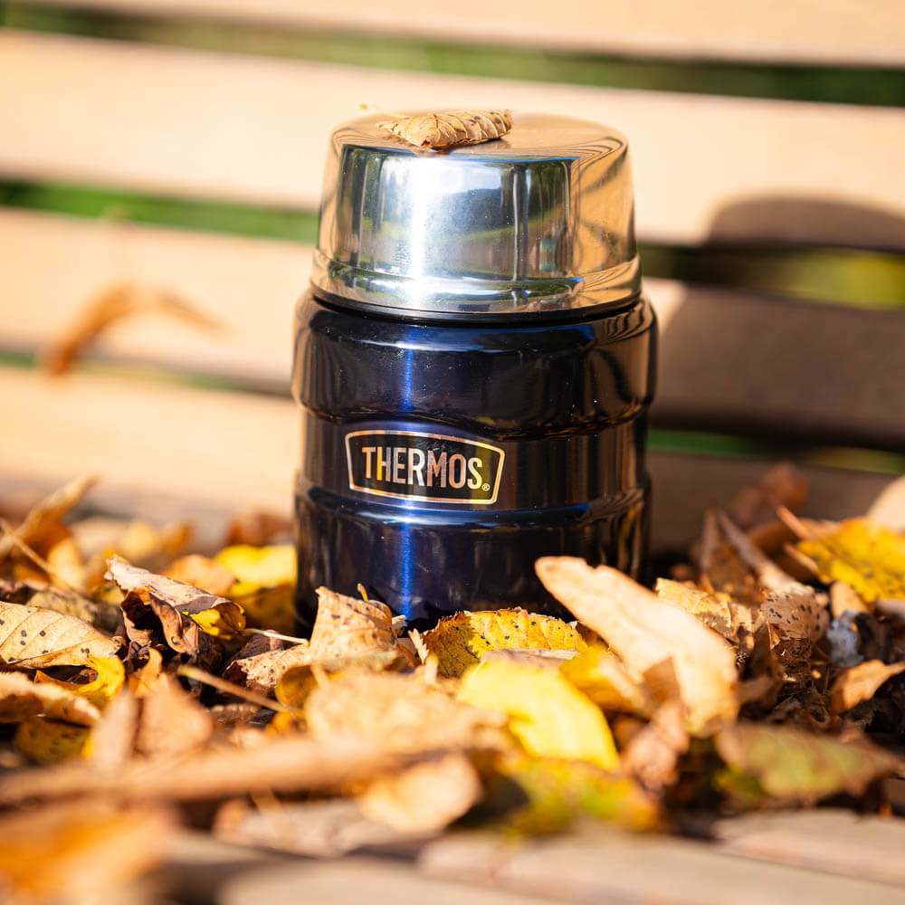 Eine blaue Thermoskanne steht auf einer Holzbank und ist von Herbstblättern umgeben. Auf dem glänzenden Deckel liegt ein kleines Blatt. Die Kanne trägt den Schriftzug "Thermos" und strahlt in der herbstlichen Umgebung.