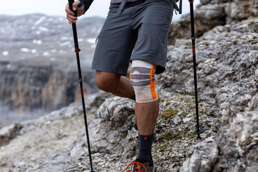 Ein Wanderer mit Trekkingstöcken und einer orangefarbenen Kniebandage geht auf einem felsigen Bergpfad. Im Hintergrund sind schroffe Felsen und neblige Berglandschaften zu sehen.