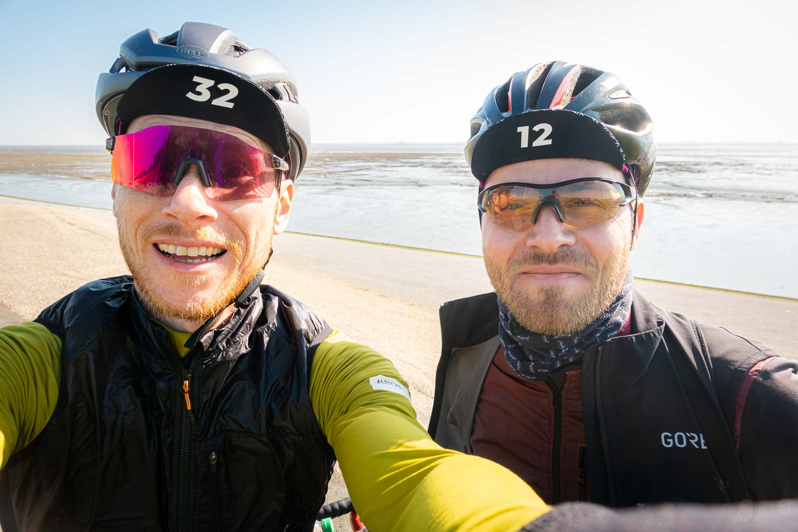 Zwei Radfahrer machen ein Selfie von sich während sie vor einem Strandpanorama stehen und lachen.